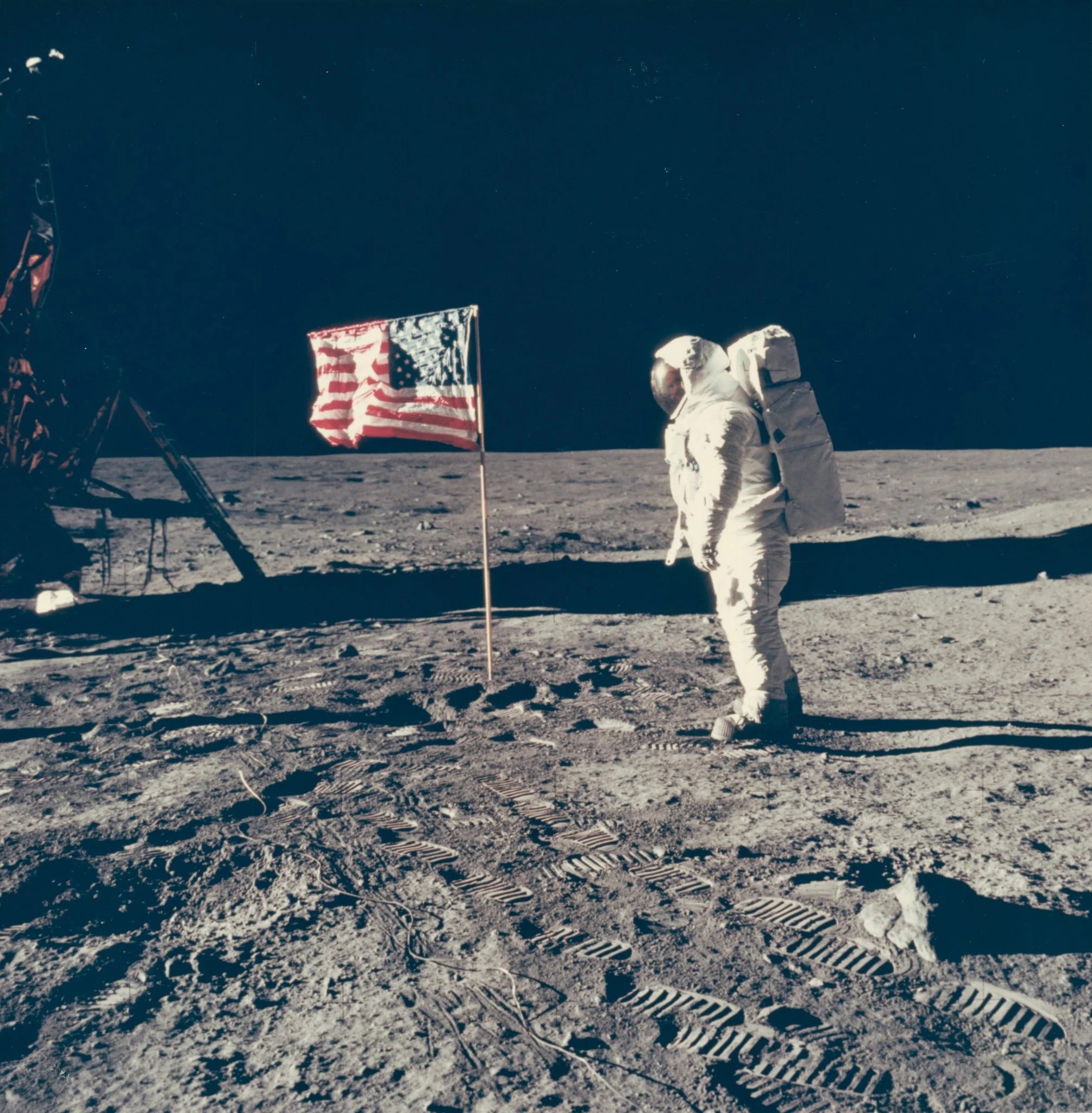 Buzz Aldrin junto a la bandera estadounidense durante Apolo 11, con la tela arrugada y sostenida por una barra horizontal sobre la superficie lunar.